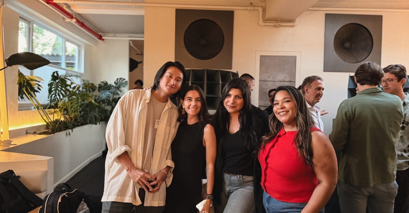 Four friends pose for a group photo at a casual indoor gathering, smiling at the camera.