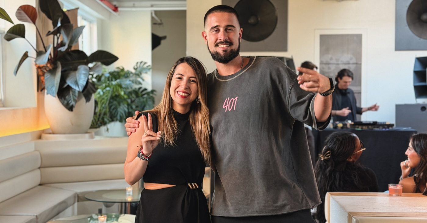 Smiling man and woman pose for a photo at a modern lounge, with a DJ and other guests in the background.