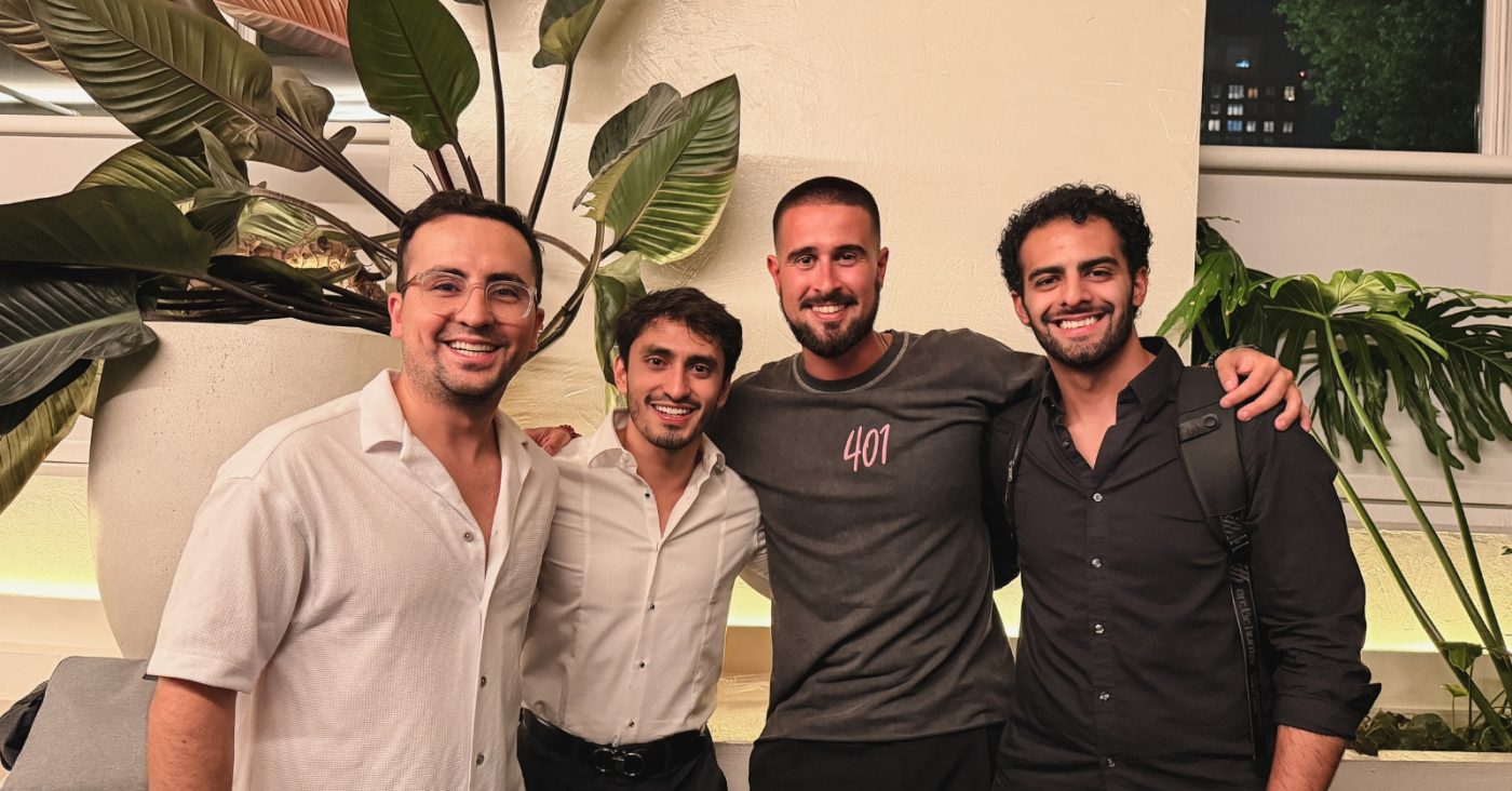 Four men posing together indoors, smiling with arms around each other, in casual to smart-casual attire, with large indoor plants in the background.