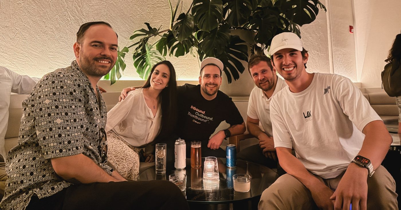 Six friends around a glass coffee table in a dim lounge, smiling at the camera with drinks and candles on the table in front of a large plant behind them.