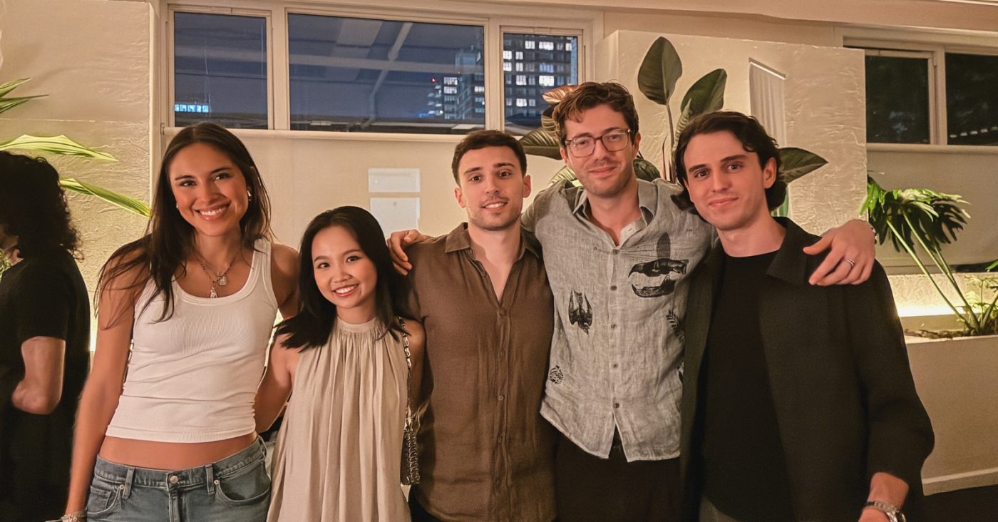 Five friends posing together indoors at a social gathering, smiling with arms around each other's shoulders.