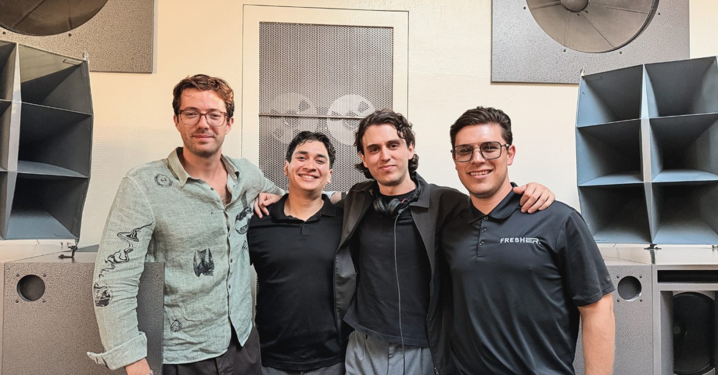 Four men stand together in a music studio with large speakers behind them, posing for a photo.