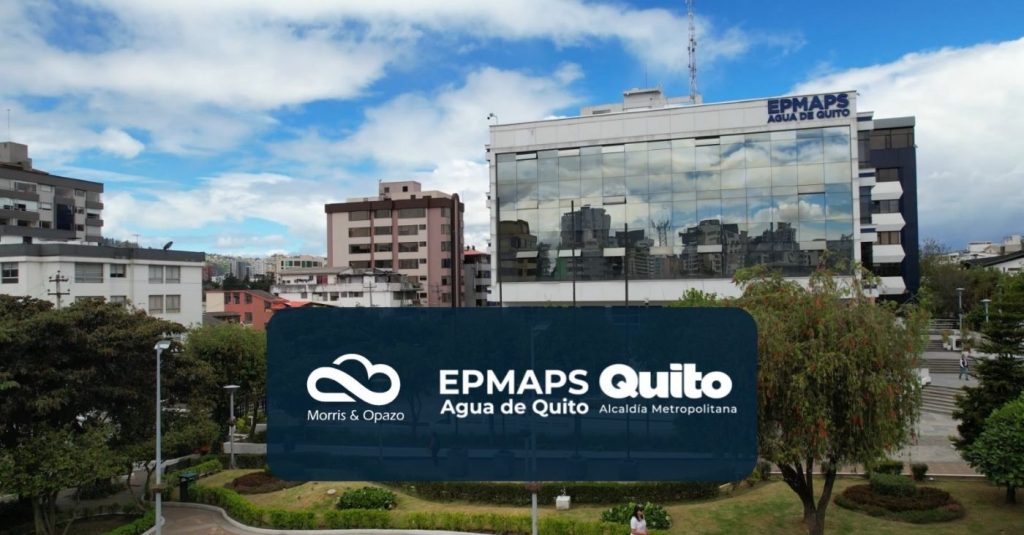 EPMaps Quito — Agua de Quito sign on a blue rounded panel in a city park, with a glass-front office building behind.